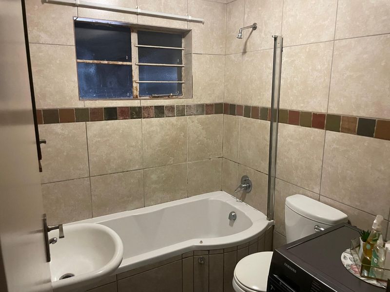 A compact bathroom featuring a bathtub with a shower attachment, glass screen, toilet, and basin, finished with neutral wall tiles.