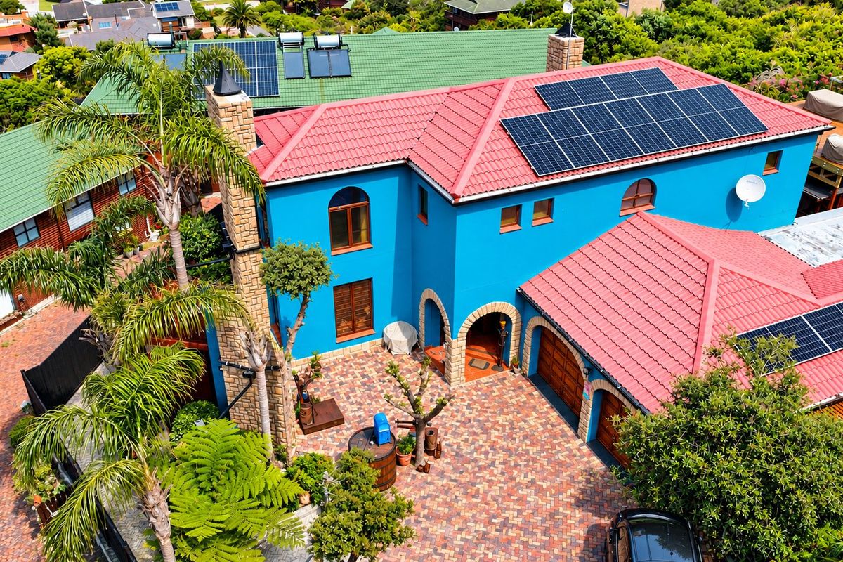 Aerial view of a vibrant blue house featuring a red tiled roof, integrated solar panels, a stone chimney, and a spacious paved courtyard entrance.