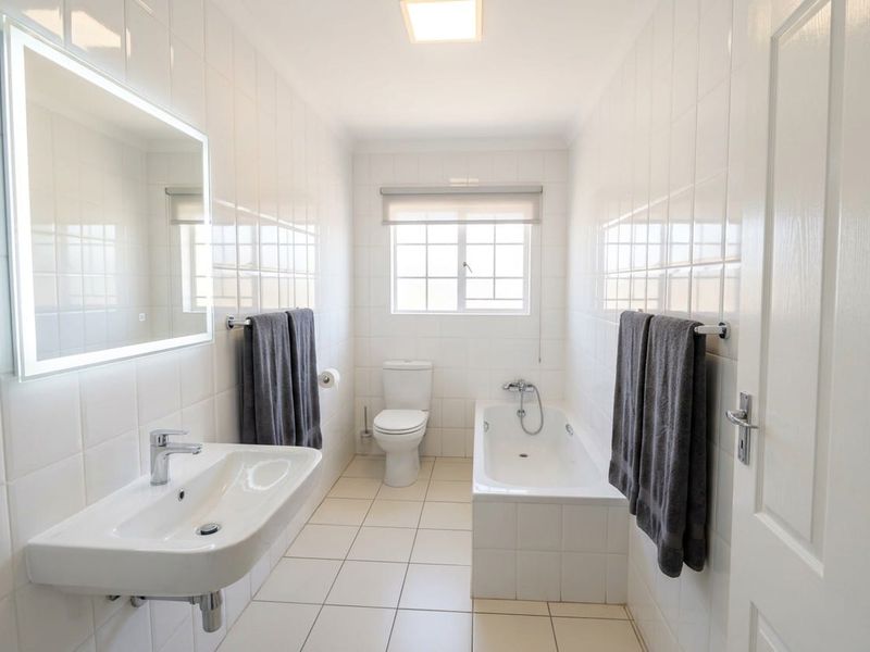 A clean, modern bathroom featuring white wall tiles, a wall-mounted basin, a built-in bathtub, and a back-to-wall toilet with an LED backlit mirror.