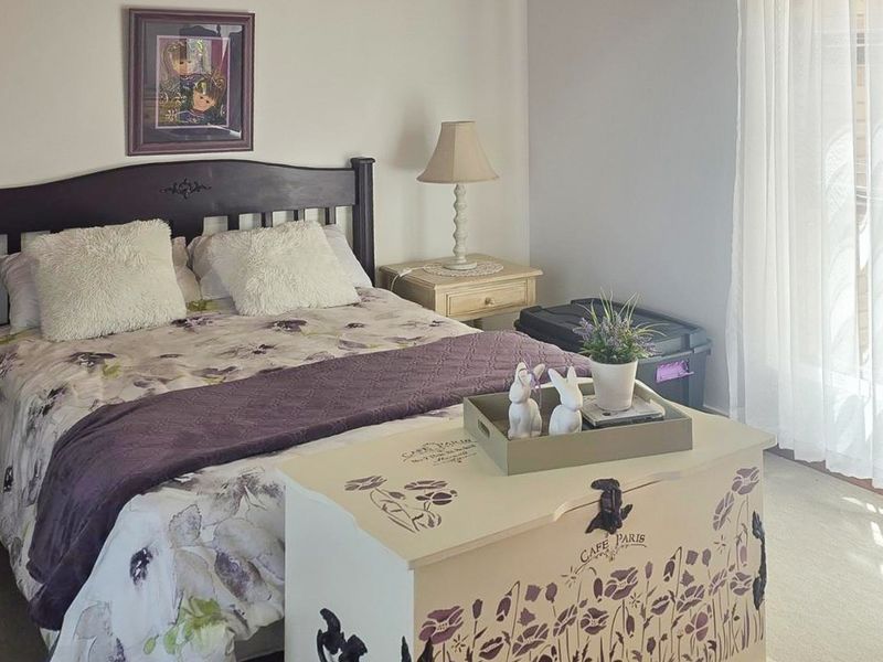 A cozy bedroom featuring neutral wall tones, carpeted flooring, and natural light provided by a large window with curtains.