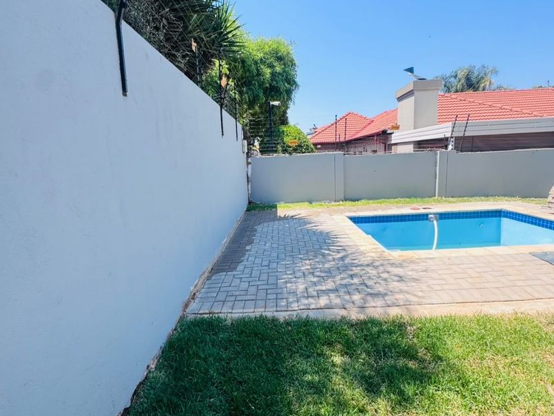 A private outdoor pool area featuring a paved deck, surrounding lawn, and secure perimeter wall with electric fencing.
