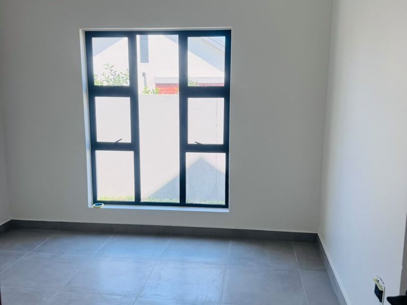 A clean, empty bedroom featuring modern grey floor tiles, neutral walls, and a large aluminium-framed window providing natural light.