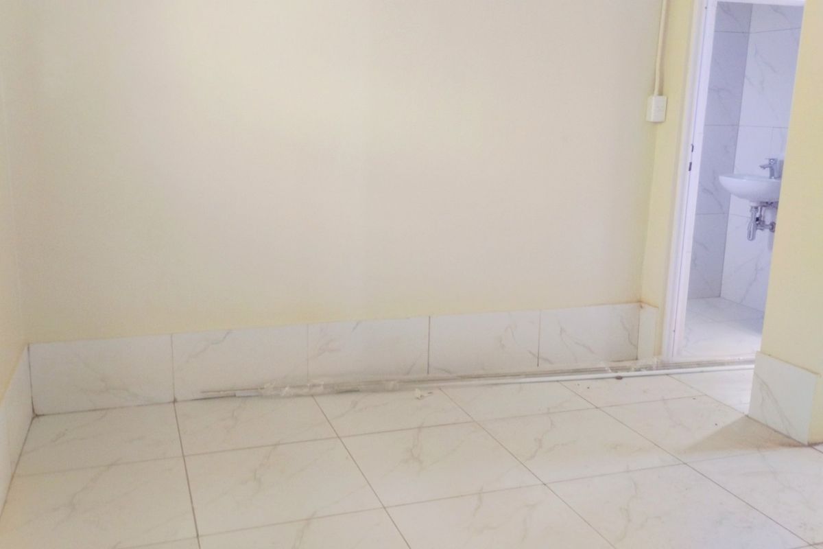 An empty bedroom featuring tiled flooring and skirting, with an adjacent bathroom visible through the doorway. The space requires finishing touches.