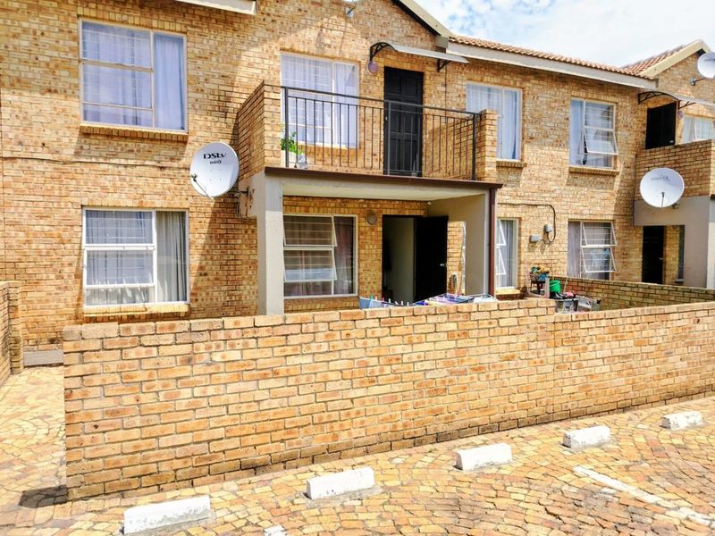 Front elevation of a face brick townhouse featuring a private balcony with wrought iron railing and a paved driveway area.