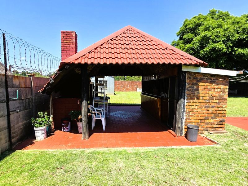 A covered outdoor patio area featuring a brick chimney and tiled roof, set within a spacious garden with perimeter security fencing.