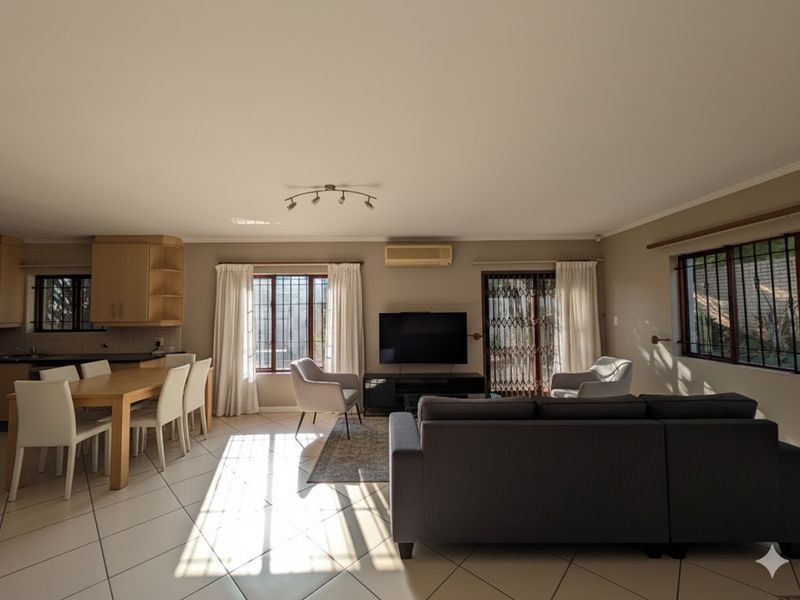 Spacious open-plan living and dining area featuring tiled floors, built-in kitchen cabinetry, and wall-mounted air conditioning, with natural light through security-fitted windows.