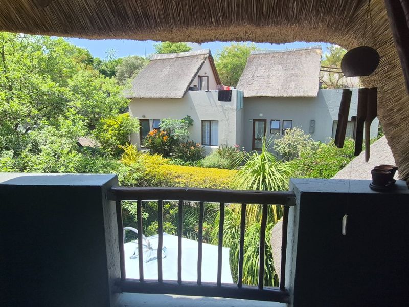 A covered balcony with a traditional thatch roof and wooden balustrade, offering a pleasant view of the surrounding garden and neighboring units.
