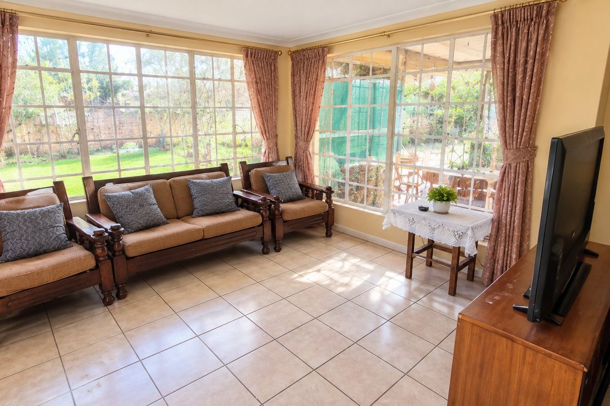 A bright living room featuring large windows with garden views and neutral ceramic floor tiles, offering a light-filled space for relaxation.