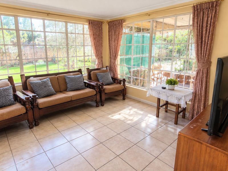 A bright living room featuring large windows with garden views and neutral ceramic floor tiles, offering a light-filled space for relaxation.