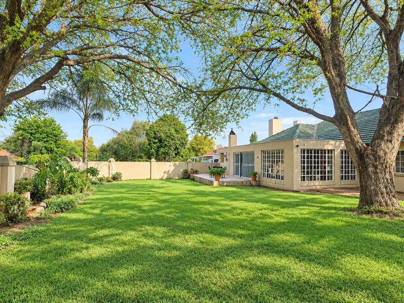 A spacious rear garden featuring a well-maintained lawn, mature trees, a brick boundary wall, and a paved patio area attached to the house.