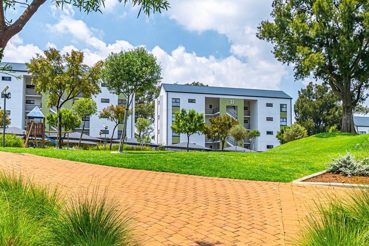 A well-maintained apartment complex exterior featuring landscaped green spaces, mature trees, and a paved walkway leading towards the residential buildings.