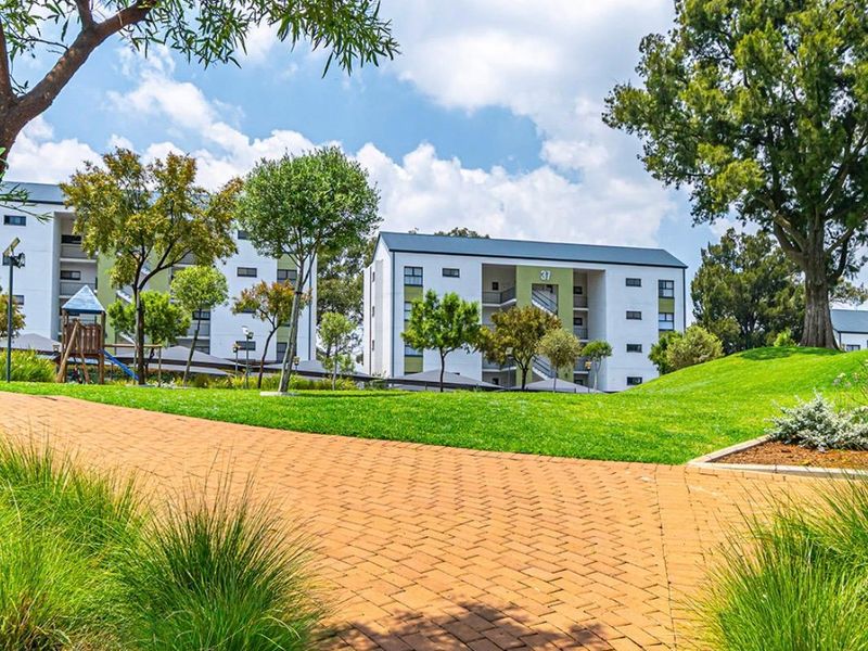 A well-maintained apartment complex exterior featuring landscaped green spaces, mature trees, and a paved walkway leading towards the residential buildings.