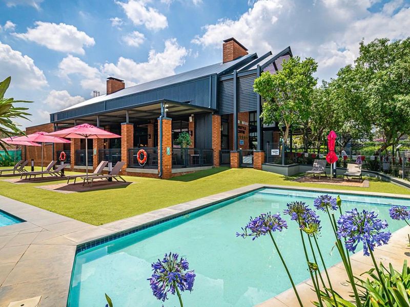 A well-maintained outdoor pool area featuring a modern building with a brick and metal facade, covered patio space, and landscaped surroundings.