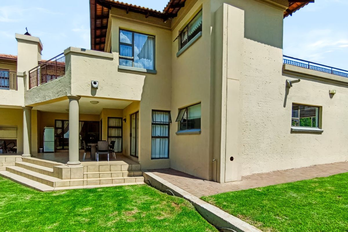 Rear view of the house featuring a covered patio, tiled terrace, built-in braai, and a well-maintained lawn with paved walkways.