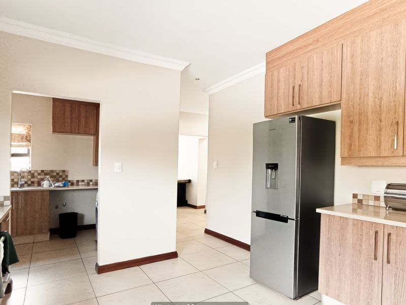 A functional kitchen featuring wood-finish cabinetry, tiled backsplash, and integrated appliances, with an adjacent scullery area and neutral floor tiling throughout.