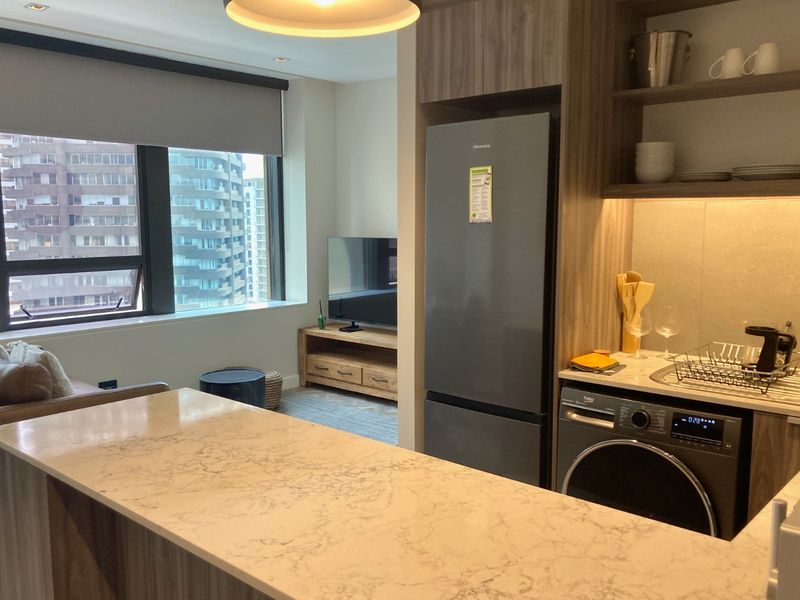 Modern open-plan apartment featuring a sleek kitchen with stone island, integrated cabinetry, and built-in shelving, seamlessly flowing into a bright living area with city views.