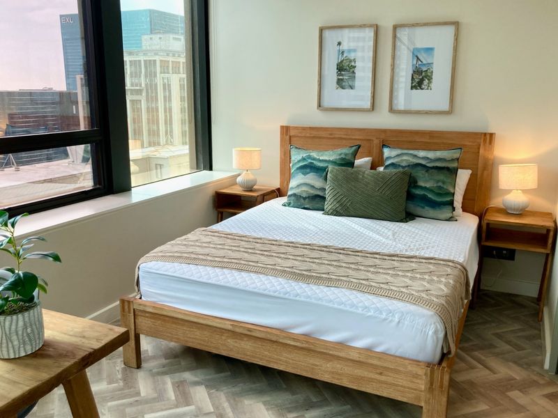 A well-lit bedroom featuring modern herringbone flooring and large floor-to-ceiling windows offering an urban city view.