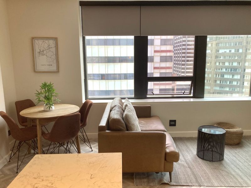 A bright open-plan living and dining area featuring modern herringbone flooring, large windows with roller blinds, and an urban city view.