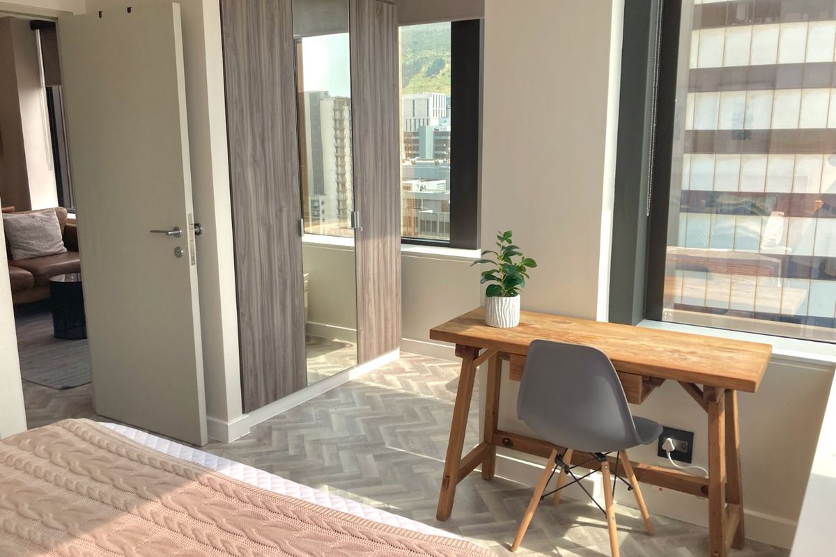 A bright bedroom featuring modern herringbone flooring, built-in wardrobes with mirrored panels, and large windows offering urban views.