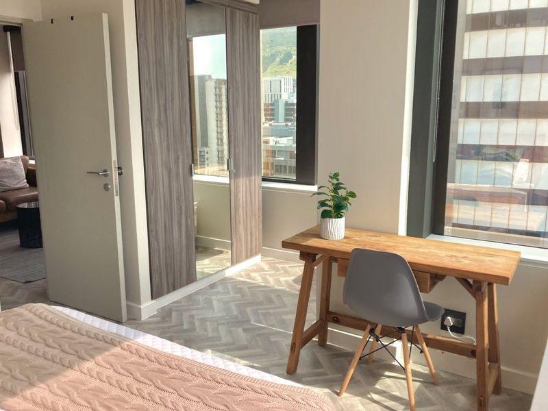 A bright bedroom featuring modern herringbone flooring, built-in wardrobes with mirrored panels, and large windows offering urban views.