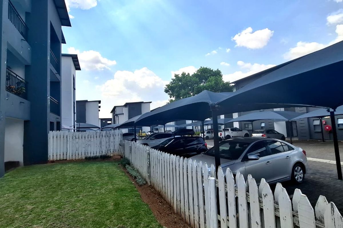 Exterior view of an apartment complex featuring a paved parking area with shade cloth carports, a lawn, and a white picket fence.