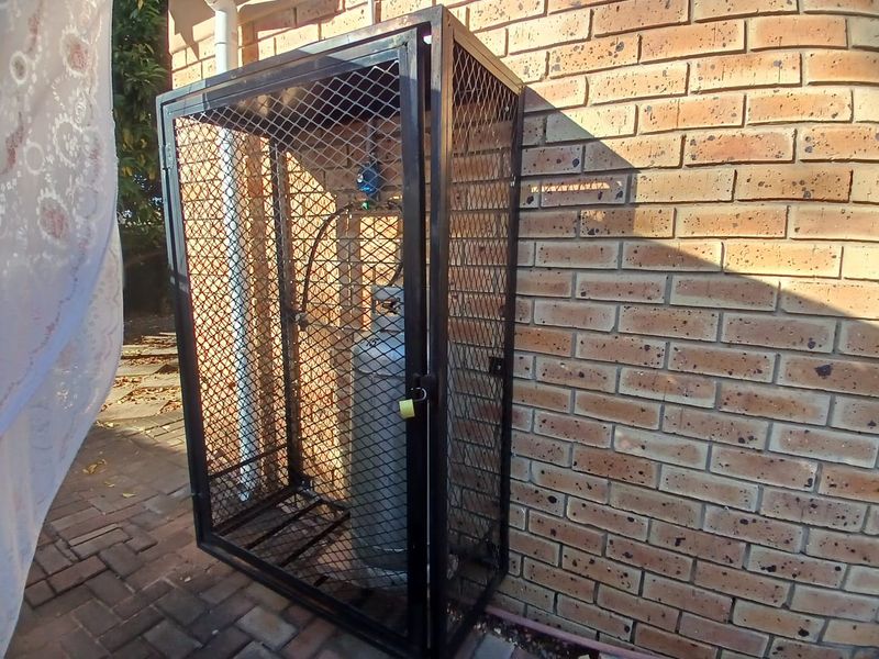 A secure metal cage enclosure mounted against an exterior brick wall, designed for safe storage of gas cylinders.