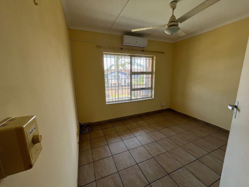 An empty bedroom featuring tiled flooring, a ceiling fan, and a wall-mounted air conditioning unit with security bars on the window.
