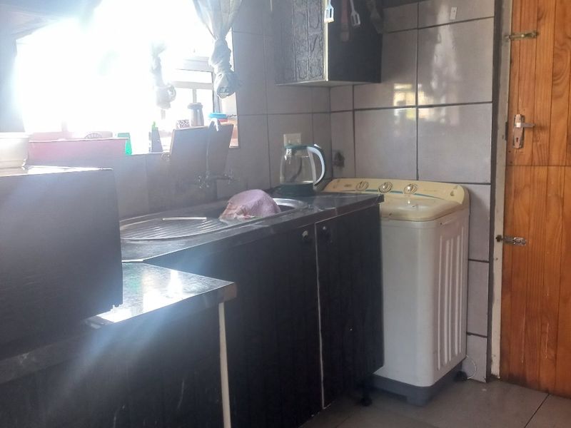 A functional kitchen area featuring built-in cabinetry, tiled splashbacks, and tiled flooring, with direct access to an adjacent room via a wooden door.