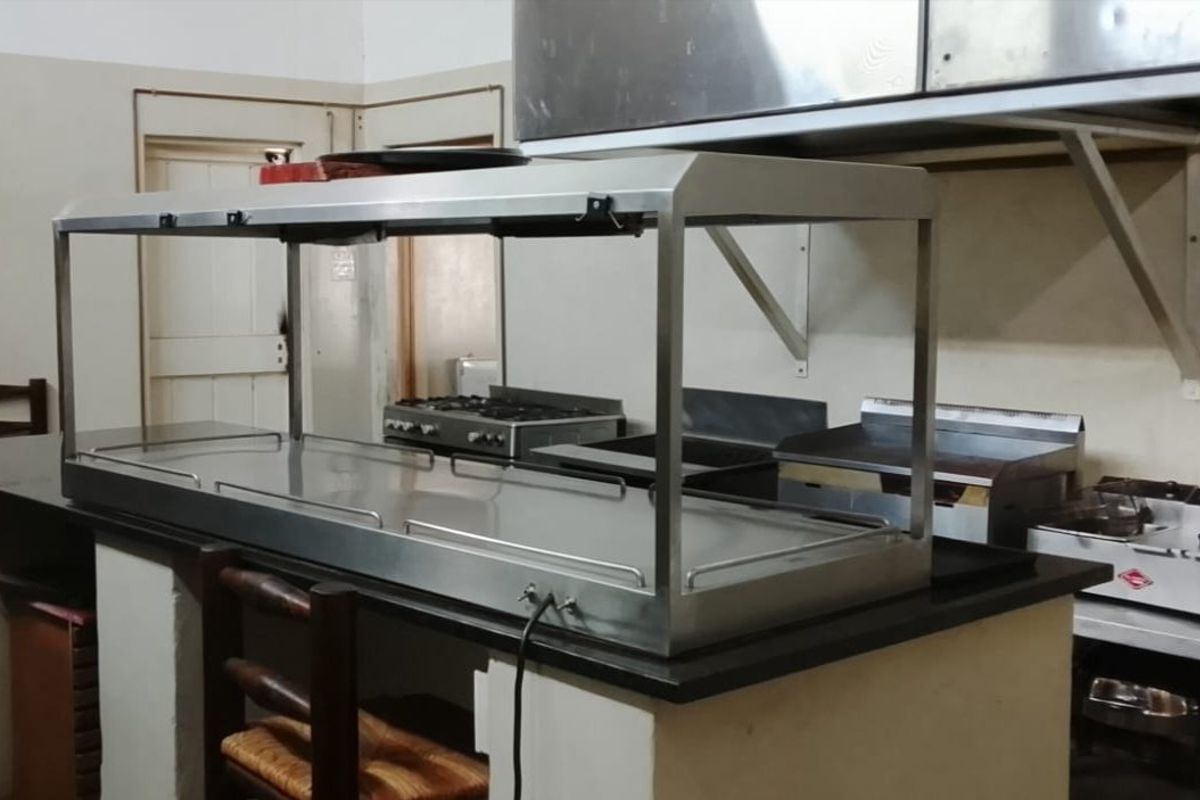 A commercial kitchen space featuring a stainless steel food warmer, gas stove, extractor hood, and various professional cooking appliances on a built-in counter.