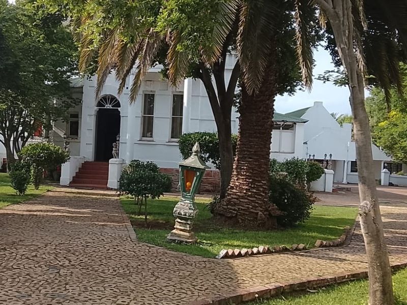 A charming commercial property entrance featuring a cobblestone driveway, mature garden landscaping, and a classic white facade with an arched doorway.
