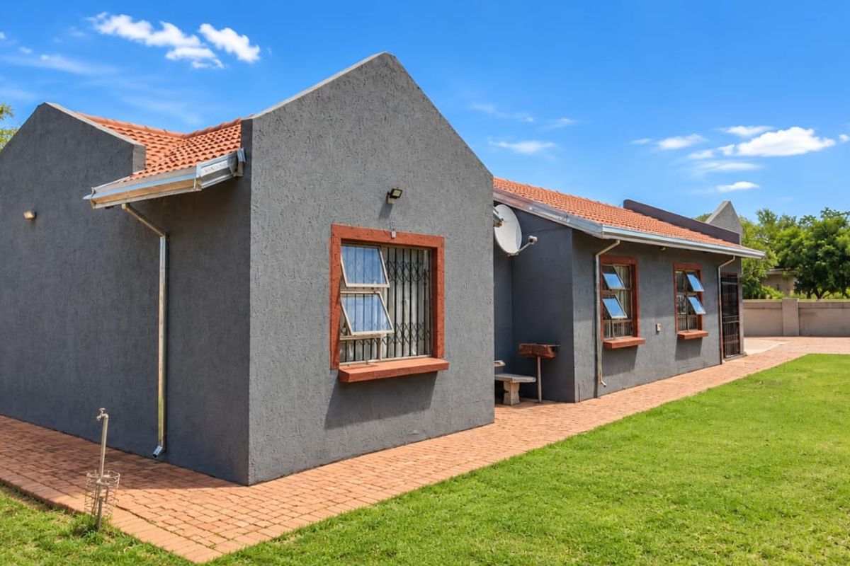 Rear view of the house featuring a grey textured facade, terracotta roof tiles, a brick paved walkway, and a well-maintained garden area.