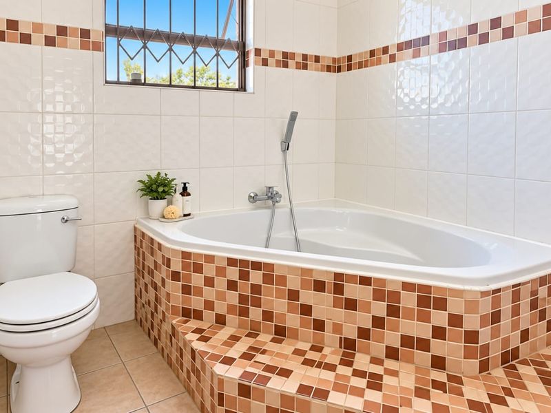 A functional bathroom featuring a built-in corner bathtub with handheld shower mixer, tiled finishes, and a window with security bars.