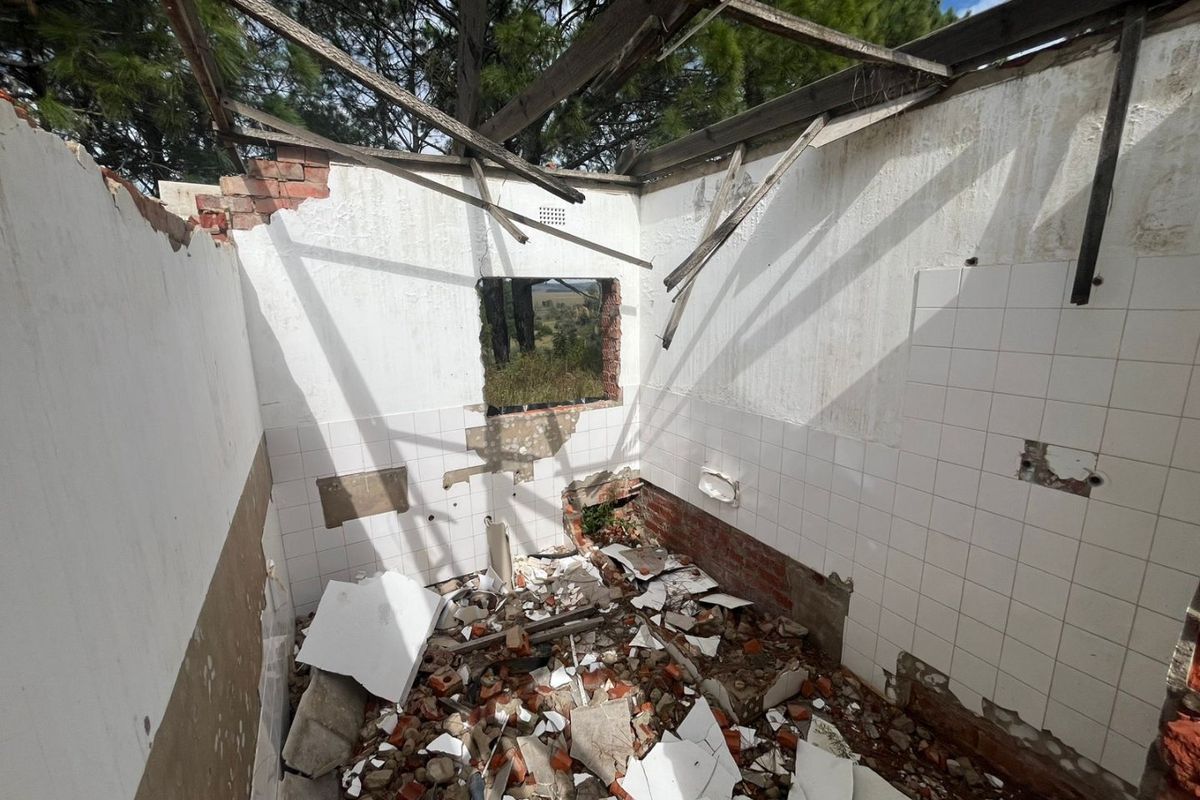 A severely dilapidated bathroom space featuring exposed brickwork, partial wall tiling, and a collapsed roof structure, requiring extensive structural renovation.