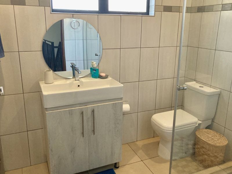 A functional bathroom featuring a modern vanity cabinet, ceramic basin, toilet, and a glass-enclosed shower with neutral wall and floor tiling.