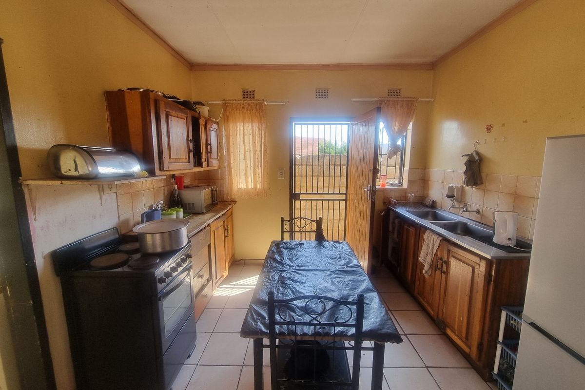 A functional kitchen featuring wooden cabinetry, double basin sink, and tiled flooring, with direct access to the rear exterior through a security gate.