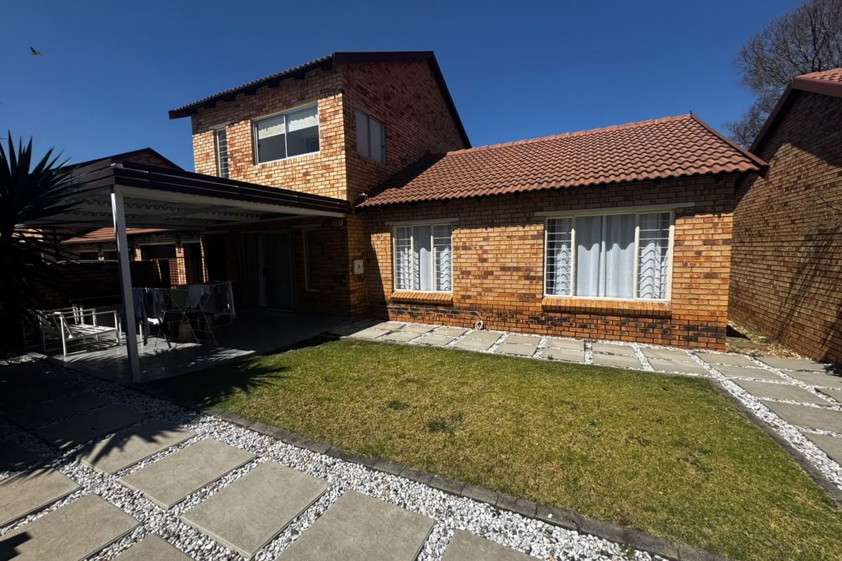 Rear view of a double-story face brick apartment featuring a covered patio, tiled roof, and a well-maintained garden with paved pathways.