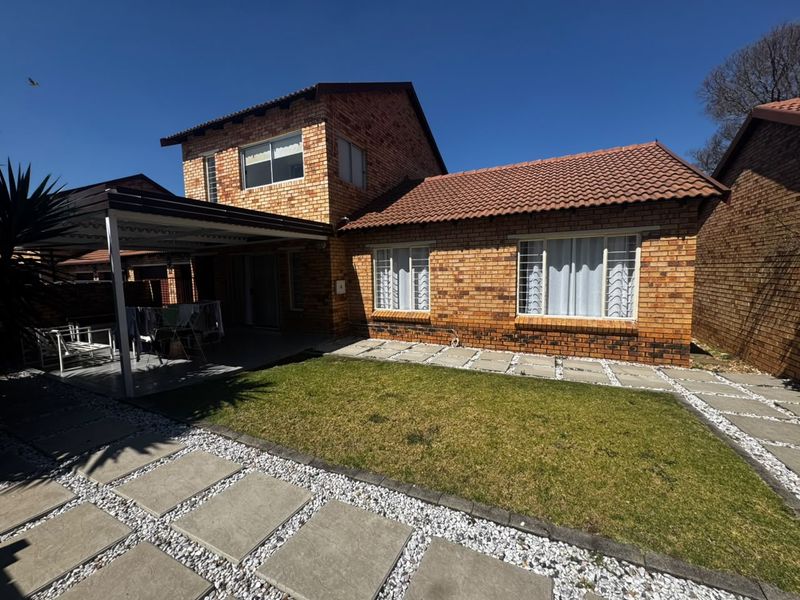 Rear view of a double-story face brick apartment featuring a covered patio, tiled roof, and a well-maintained garden with paved pathways.