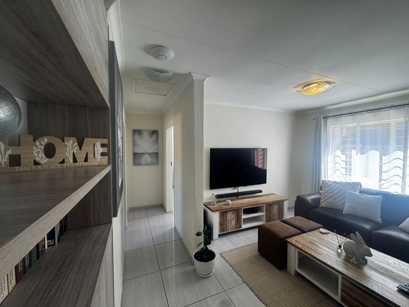 A well-maintained living room featuring neutral tiled flooring, clean wall finishes, and natural light through a window with security bars.