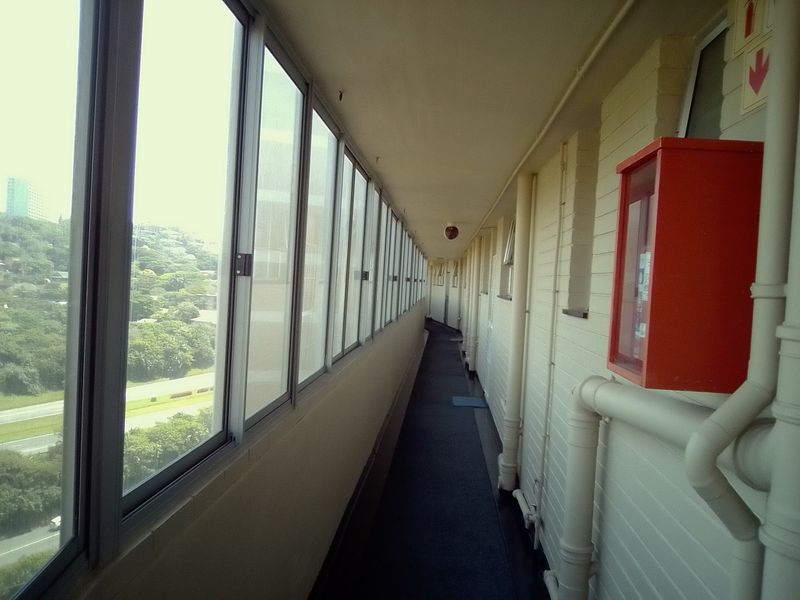 A long apartment building corridor featuring large windows, exposed plumbing, and a wall-mounted fire hose reel.