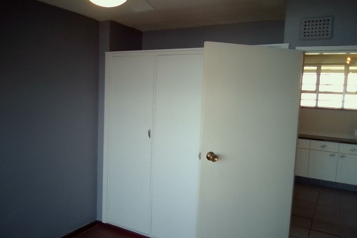 A vacant bedroom featuring a built-in wardrobe, neutral wall paint, and a view into the adjacent kitchen area.