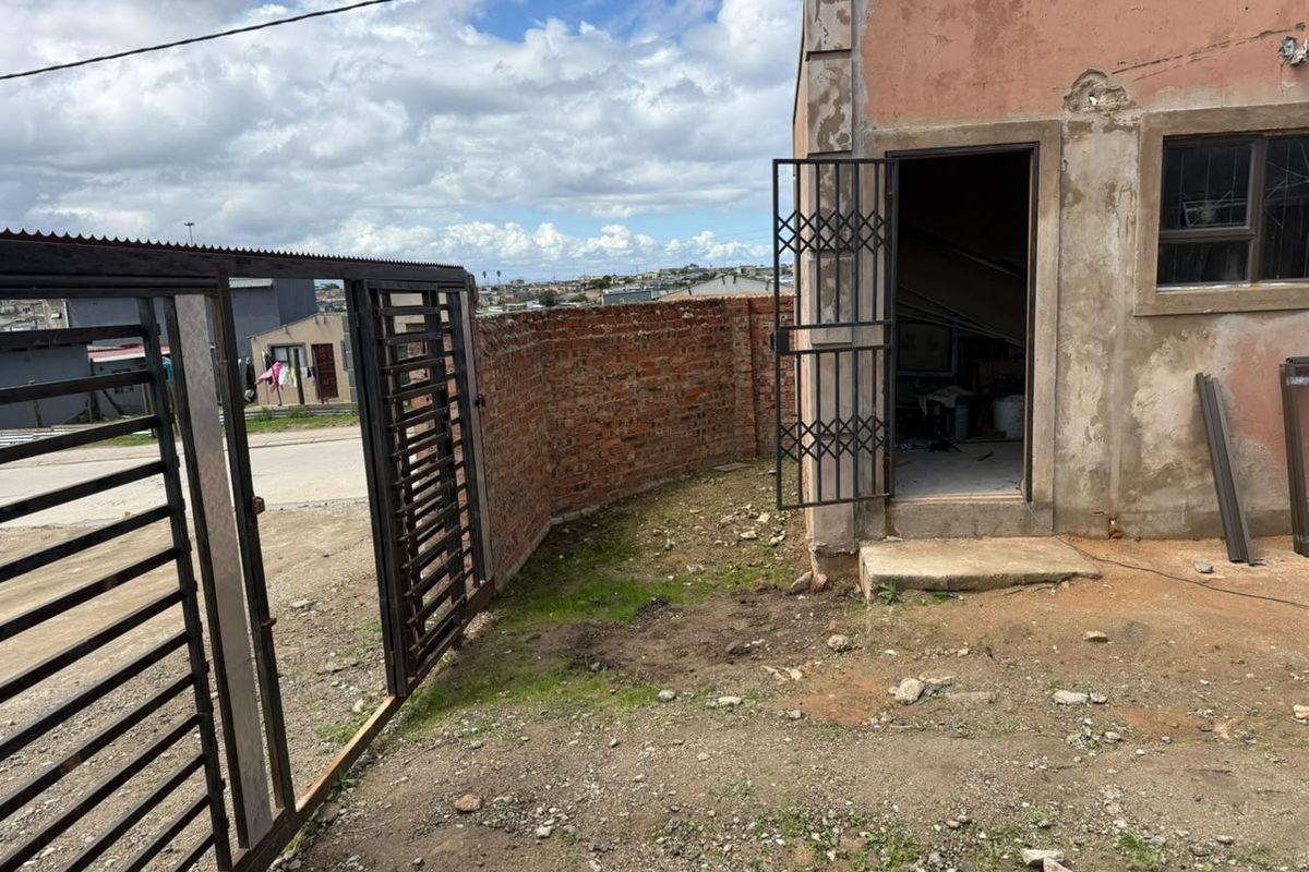 Exterior view of a property featuring a metal security gate, brick boundary wall, and a building with weathered exterior finishes and security door access.