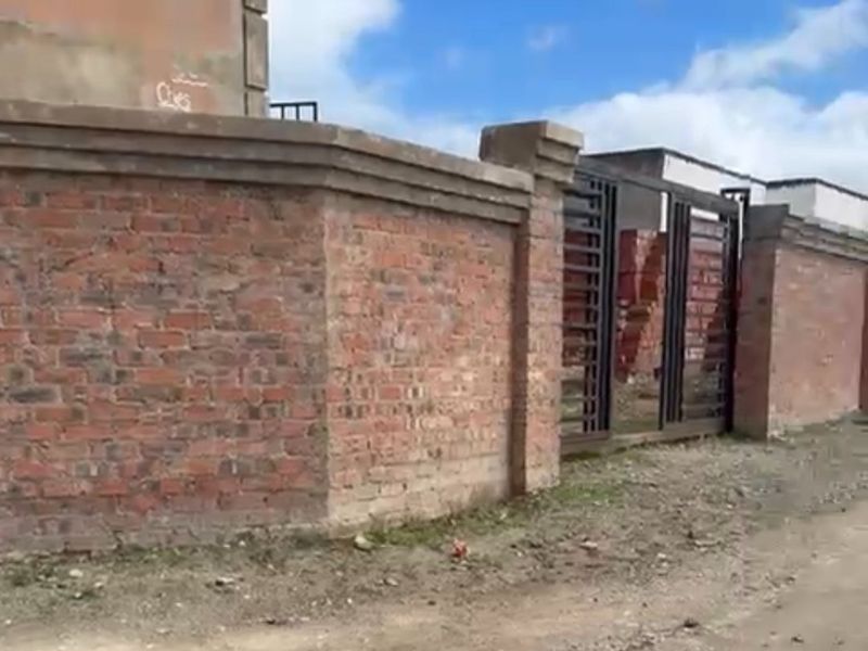 Front view of the property featuring a brick boundary wall and metal gate, situated on an unpaved road.