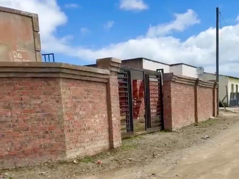 Exterior view of a property featuring a brick boundary wall and metal gate, situated along an unpaved road.