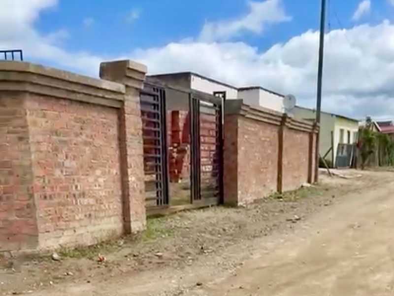 Street view of a property featuring a brick boundary wall and metal driveway gate, situated along an unpaved road.