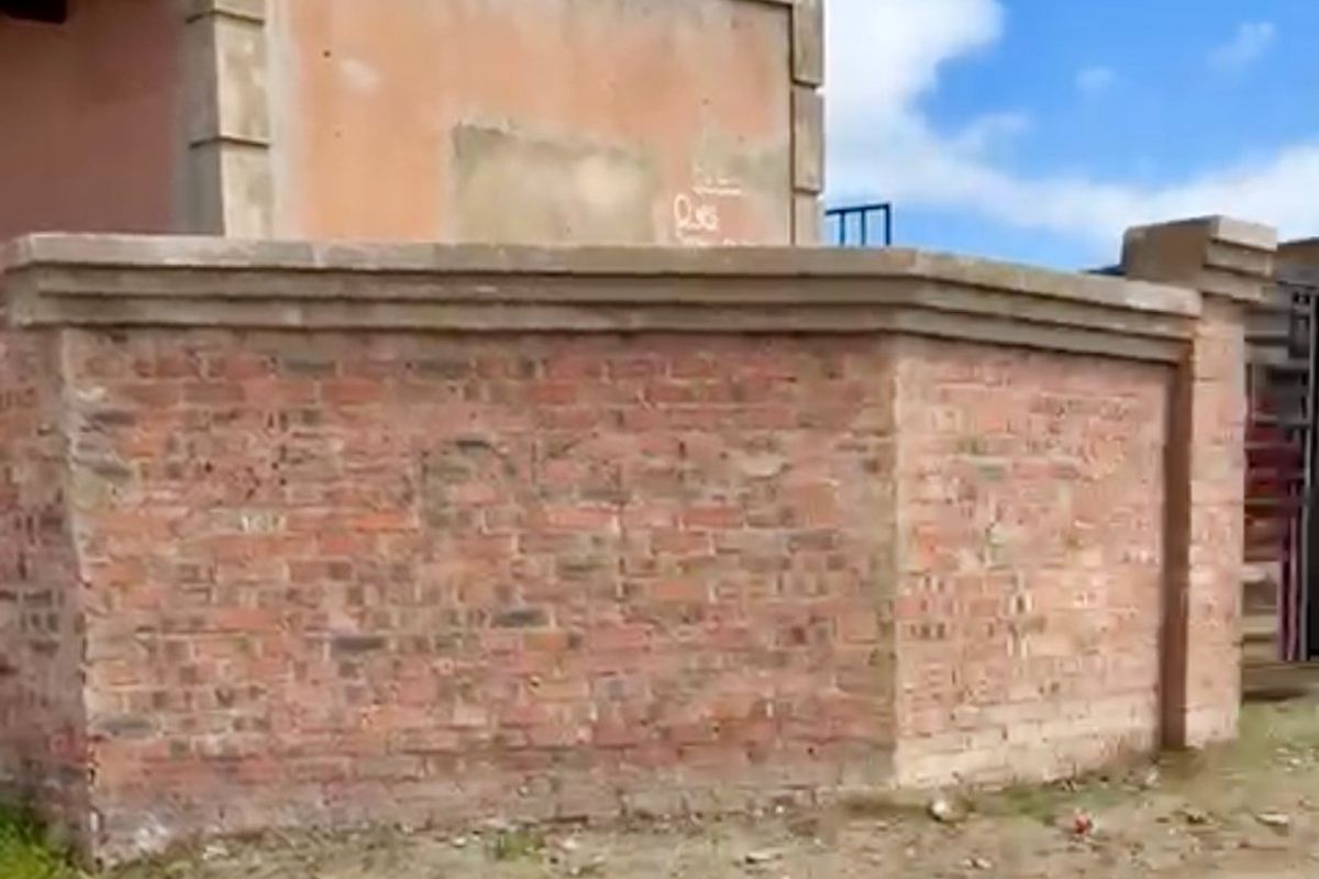 Exterior view of a property featuring a brick boundary wall and an unfinished plastered masonry structure on an unpaved lot.