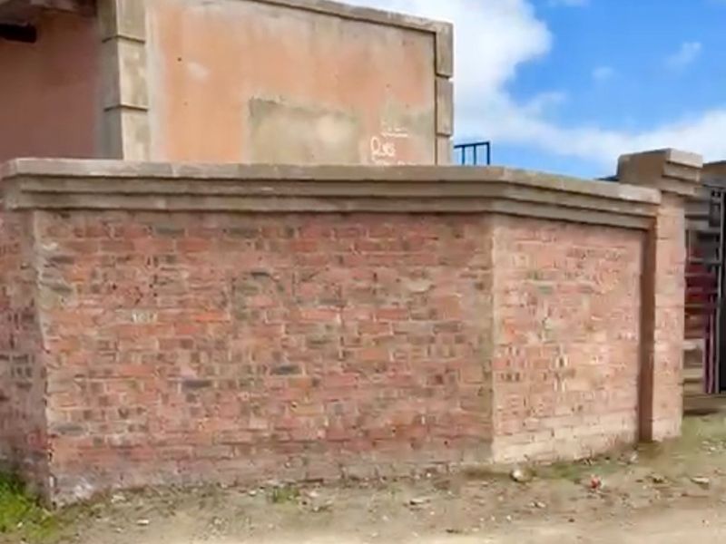 Exterior view of a property featuring a brick boundary wall and an unfinished plastered masonry structure on an unpaved lot.