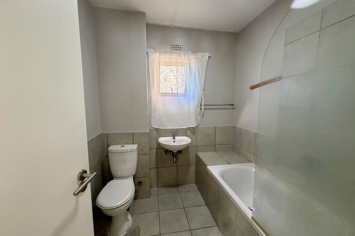 A functional bathroom featuring a built-in bathtub with a glass screen, a wall-mounted basin, and a standard toilet with neutral floor and wall tiling.