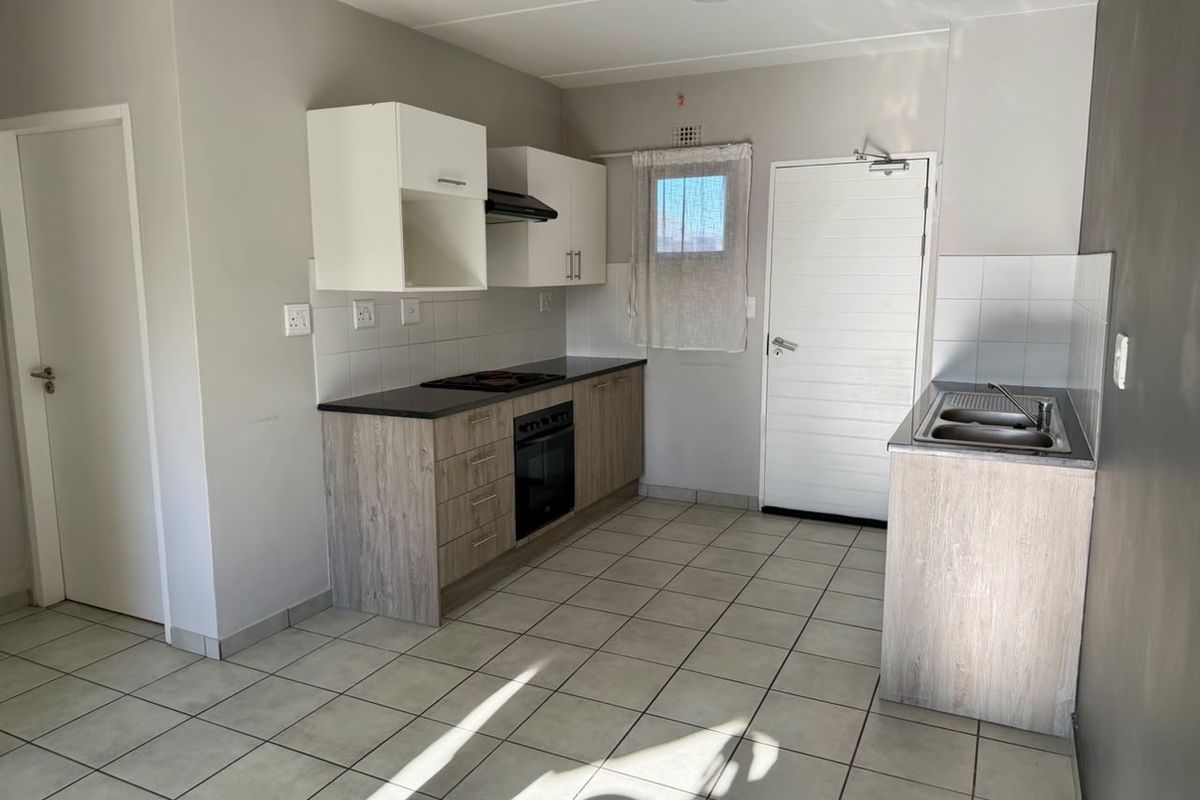 A clean, functional kitchen featuring light wood-look cabinetry, dark countertops, built-in oven and hob, and neutral tiled flooring.