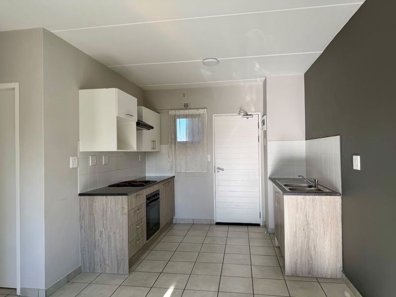 A clean, functional kitchen featuring light wood-look cabinetry, dark countertops, built-in oven and hob, and tiled flooring in an open-plan layout.