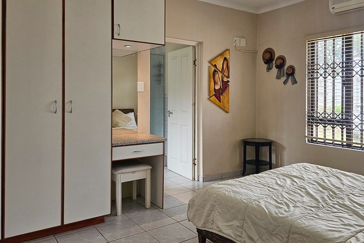 A functional bedroom featuring built-in cupboards, a vanity area, tiled flooring, and an air conditioning unit, with security bars on the window.